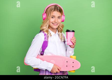 Ritratto di bella ragazza allegra che ascolta melodia bere latte tenendo skate isolato su sfondo di colore verde Foto Stock