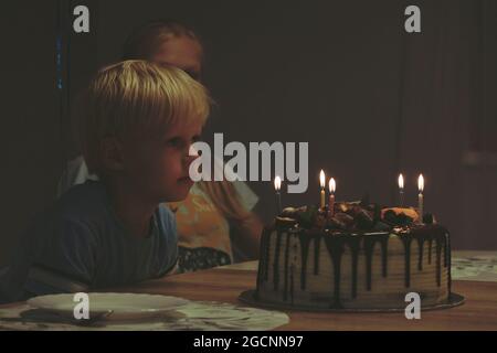 I bambini di una festa di compleanno si siedono ad un tavolo in una stanza buia e guardano una torta di compleanno con candele brucianti. Atmosfera di casa festosa. Foto Stock