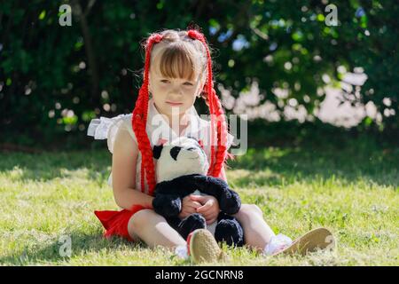 La bambina con i pigtail rossi si siede sull'erba del parco con un panda di orso giocattolo. Foto di alta qualità Foto Stock