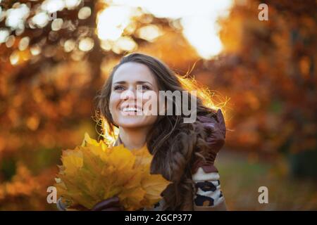 Ciao ottobre. Donna trendy sorridente di 40 anni in camice beige con foglie gialle d'autunno fuori sul parco cittadino in autunno. Foto Stock