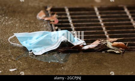 Un primo piano di una maschera protettiva personale usata scartata che entra in una griglia metallica su uno scarico. Crescente preoccupazione ambientale per quanto riguarda lo smaltimento sicuro Foto Stock