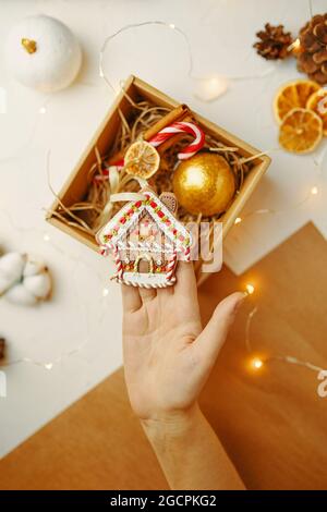 Vista dall'alto della casa di pan di zenzero natalizio in mano. Confezione regalo con decorazioni natalizie. Carta da imballaggio artigianale, coni, garland e arance secche su tavola bianca. Atmosfera di Capodanno. Per cartoline. Foto Stock