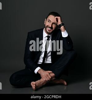 Un uomo sorridente, adulto, porticato in tuta da ufficio nera, si siede a barefooted sul pavimento con gambe incrociate che tengono la mano alla testa Foto Stock