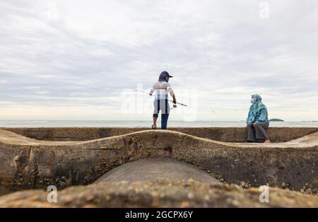 Port Dickson, Malesia - 15 dicembre 2020: Giovane uomo e sua moglie che pescano in un frangiflutti sulla spiaggia. L'angler getta la sua canna da pesca o l'angelo Foto Stock