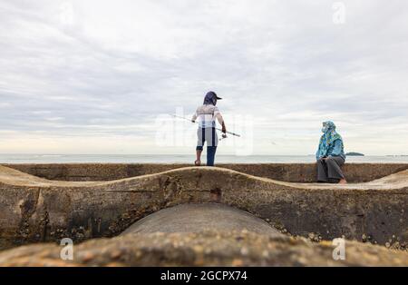 Port Dickson, Malesia - 15 dicembre 2020: Giovane uomo e sua moglie che pescano in un frangiflutti sulla spiaggia. L'angler getta la sua canna da pesca o l'angelo nel Foto Stock