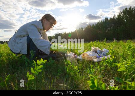 Giovane donna in guanti di gomma sulla natura raccoglie i rifiuti di plastica in una giornata estiva di sole sullo sfondo degli alberi. Protezione ambientale. Selez Foto Stock