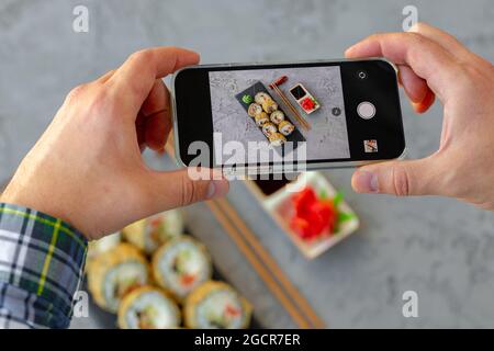 Mani maschili che tengono lo smartphone e scattano una foto di sushi roll su sfondo grigio Foto Stock