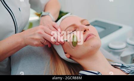Primo piano di massaggio viso con un rullo di giada sul viso della donna presso il salone di bellezza. Cosmetologa che fa la procedura per la giovane donna. Foto Stock