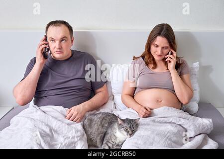 Un uomo e una donna incinta stanno parlando sui loro telefoni a letto Foto Stock