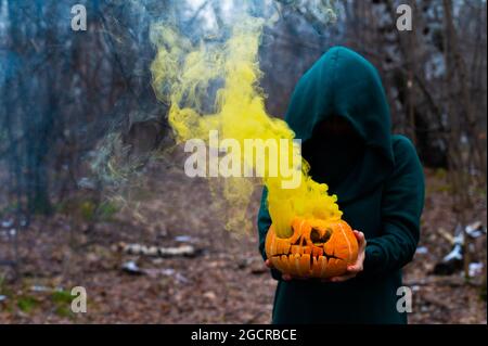 Una strega creepy tiene una zucca fumante in una foresta profonda. Jack o lanterna emette fumo giallo per Halloween Foto Stock