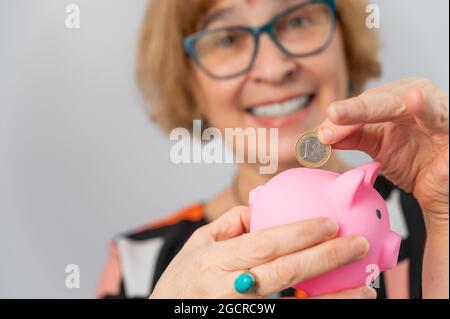 Una donna anziana con gli occhiali mette una moneta in una banca piggy su uno sfondo bianco Foto Stock