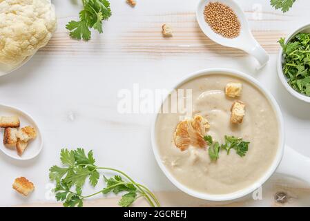 Zuppa di crema di cavolfiori con erbe verdi fresche e crostini in ciotola bianca in ceramica su tavola di legno bianco e beige. Piatto posare con piatto pronto per mangiare e. Foto Stock