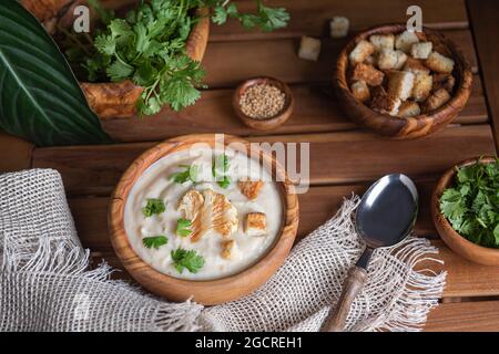 Zuppa di crema di cavolfiori fatta in casa con erbe e crostini appena tritati in ciotole di legno su tavola di legno marrone. Tovagliolo di lino, cucchiaio, semi di senape e. Foto Stock