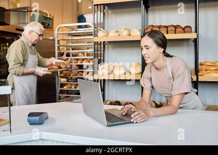 Giovane donna fornaio che usa un computer portatile contro il suo collega maschio maturo che porta pane fresco Foto Stock