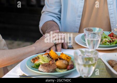 Mani di coppia affettuosa matura seduti al tavolo servito e cena Foto Stock