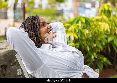 Donna adulta che si rilassa all'esterno appoggiandosi su una sedia con le mani attaccate dietro il collo e guardandosi in alto. Donna afro-americana che si gode la giornata. Foto Stock