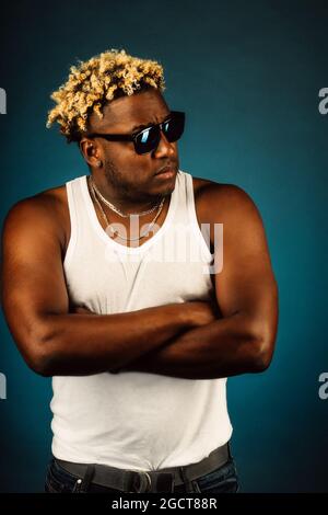 Questo è un ritratto da studio di un afroamericano in occhiali da sole e una T-shirt bianca su uno sfondo blu scuro. Un ragazzo sciuto con una pettinatura alla moda con i pigtail tiene le mani sul petto e si guarda via. Foto Stock