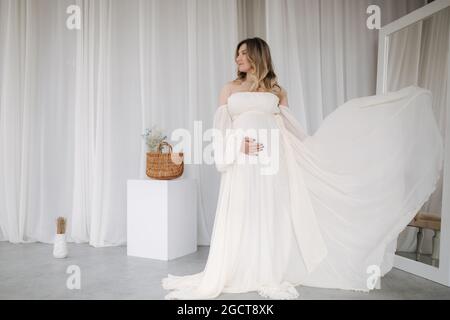 Bella donna incinta in elegante abito bianco in posa per fotografa in studio. Sfondo di tulle bianco Foto Stock