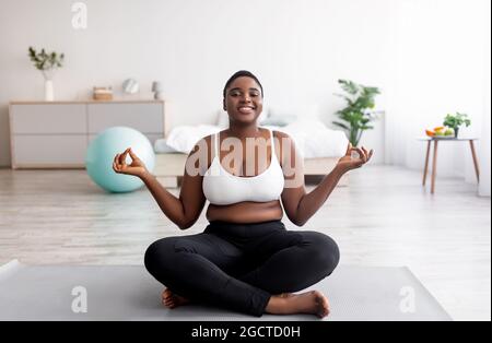 Donna nera sovrappeso seduta sul tappeto sportivo in posa lotus, meditando, facendo pratica yoga a casa Foto Stock