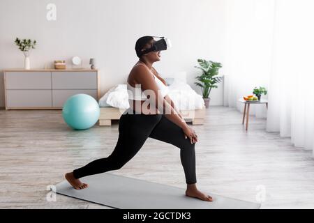 Vista laterale di una donna nera in sovrappeso in un visore VR con l'app per la realtà virtuale, che fa affondo a casa Foto Stock