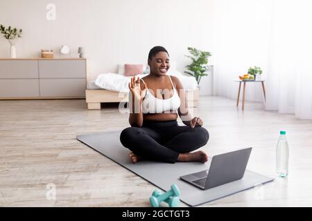 Donna nera in sovrappeso seduta sul tappetino yoga di fronte al computer portatile, saluto personal trainer, agitando alla webcam, spazio per la copia Foto Stock