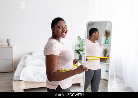 Donna curvy afro che misura la vita con nastro vicino a specchio, soddisfatto con il risultato di perdita di peso, ottenendo il successo dimagrante Foto Stock