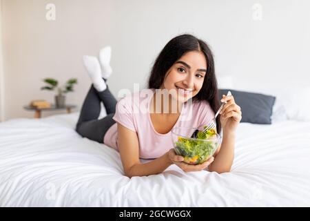 Mangiare sano, mangiare e disintossicare concetto. Bella donna indiana con insalata veggie fresca, sdraiata a letto a casa Foto Stock