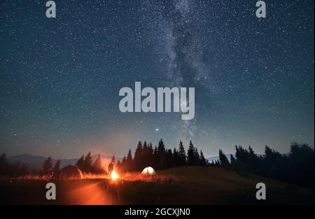 Silhouette di viaggiatore maschile in piedi vicino al falò e tende turistiche sotto il bel cielo notturno con stelle e Via Lattea. Magnifica vista della stella blu Foto Stock