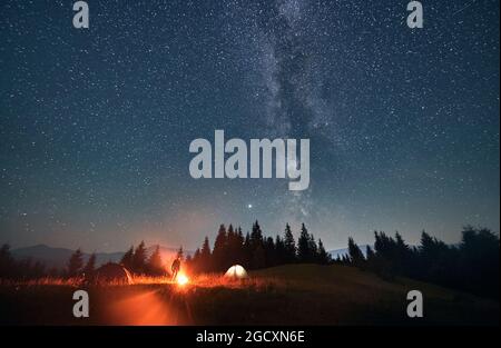 Silhouette di viaggiatore maschile in piedi vicino al falò e tende turistiche sotto il bel cielo notturno con stelle e Via Lattea. Magnifica vista della stella blu Foto Stock