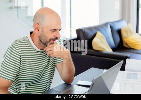 Uomo di mezza età con t-shirt a righe che lavora da casa con il suo laptop in soggiorno Foto Stock