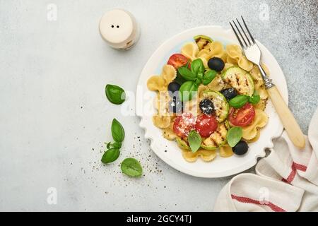 Insalata di pasta fiocchi con verdure zucchine grigliate, pomodori ciliegini, olive, basilico e parmigiano in piatto bianco su ardesia leggera, pietra o calcestruzzo Foto Stock