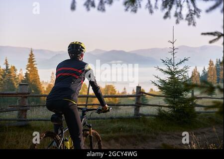 Vista dal lato posteriore corto del ciclista maschile in bici da ciclismo con colline sullo sfondo. Uomo in bicicletta indossando casco di sicurezza mentre godendo la bicicletta sbarazzarsi Foto Stock