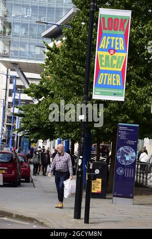 Manchester, Regno Unito, 10 agosto 2021. La gente passa accanto a un cartello locale che li invita a 'guardarsi l'un l'altro a Manchester, Regno Unito. Covid-19 o il tasso di infezione del coronavirus cade a Manchester ma aumentano in altri tre distretti più grandi di Manchester: Stockport, Bury e Trafford, secondo i dati più recenti dalla sanità pubblica Inghilterra. Credit: Terry Waller/Alamy Live News Foto Stock