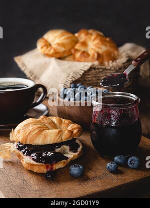 Croissant con confettura di mirtilli e mirtilli su tavola di legno con caffè, ciotola di frutti di bosco e cesto di croissant sullo sfondo Foto Stock