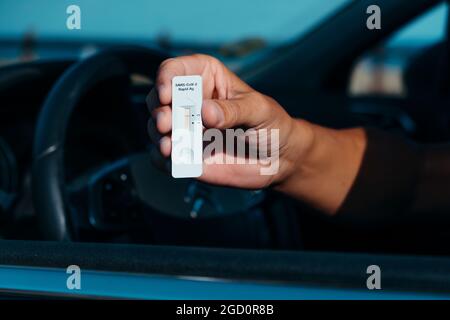 un giovane caucasico, seduto al posto di guida della sua auto, mostra un covid-19 dispositivo di prova con un risultato negativo Foto Stock