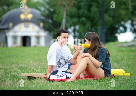 Due ragazze adolescenti che mangiano orecchie di mais bollito insieme seduti sull'erba di un prato, parco cittadino. 7 agosto 2021. Kiev, Ucraina Foto Stock