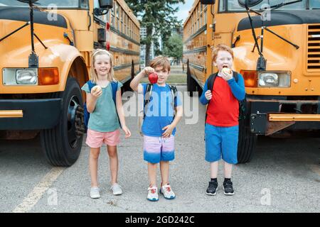 Bambini ragazzi e ragazze studenti amici mangiare mele snack sano da scuola bus giallo all'aperto. Istruzione e ritorno a scuola nel mese di settembre. Foto Stock