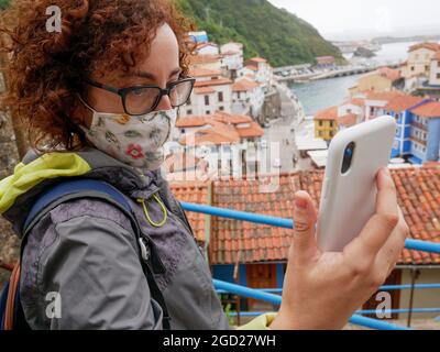 Una donna che indossa una maschera facciale usa il suo telefono cellulare per prendere un selfie in un villaggio Foto Stock