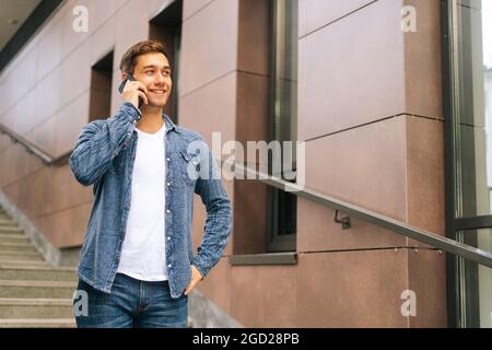 Ritratto di medio scatto di un bel giovane sicuro che parla sul telefono cellulare in piedi sulle scale del moderno edificio dell'ufficio Foto Stock