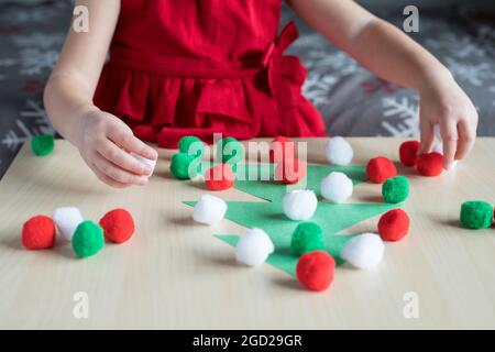 Vacanze di Natale bambini attività artigianali a casa. Piccola ragazza che gioca carta decorazione albero di Natale con piccoli pomposs. Foto Stock