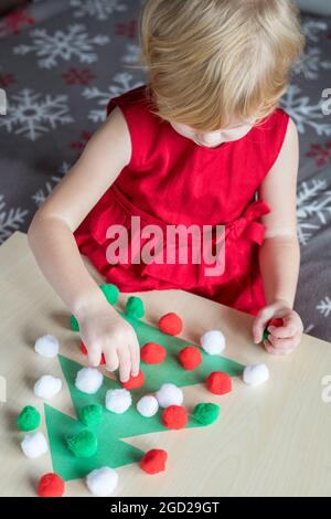 Vacanze di Natale bambini attività artigianali a casa. Ragazza piccola in vestito rosso che gioca carta decorazione albero di Natale con piccoli pomposs. Foto Stock