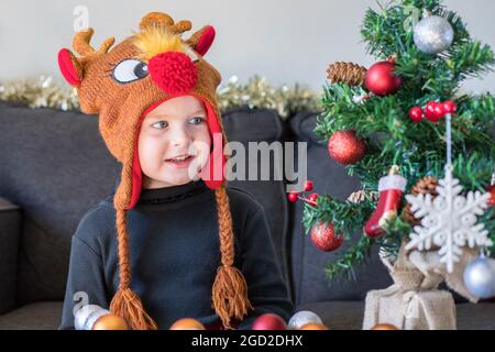 Concetto di Natale e Capodanno. Bambino sorridente seduto sul divano e guardando l'albero di Natale artificiale decorato con palle e fiaccello di neve Foto Stock