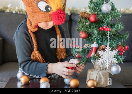 Concetto di preparazione per le festività di Natale e Capodanno. Bambino decorazione albero di Natale con palle e fiocchi di neve indossare abito e cappello di santa. Abete a Foto Stock