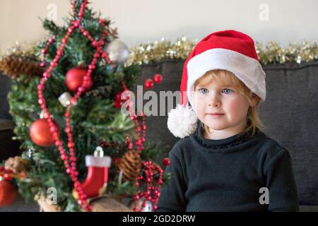 Concetto di preparazione per le festività di Natale e Capodanno. Felice bambino sorridente che decora e guarda l'albero di Natale, indossando abiti e cappello di santa. Foto Stock