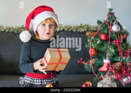 Bambino con regalo di natale. Festa di Natale e Capodanno. Bambino che tiene la scatola del regalo con sorpresa, che indossa l'abito e il cappello di santa, decorato artificio Foto Stock