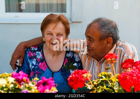 Coppie anziane che abbracciano nel giorno d'estate. Gioiosa bella coppia anziana di marito e moglie sorridenti e in posa su fiori giardino sfondo. Amore e famiglia Foto Stock