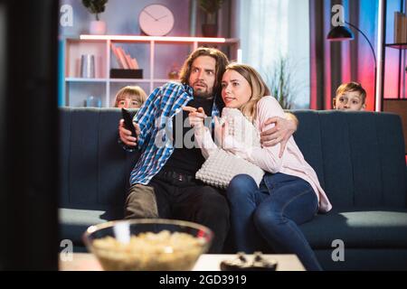 Shared bearded marito abbracciare la sua moglie spaventata durante, guardando film horror in TV a casa in serata. Due bambini spaventati sono nascosti dalla paura dietro il divano Foto Stock