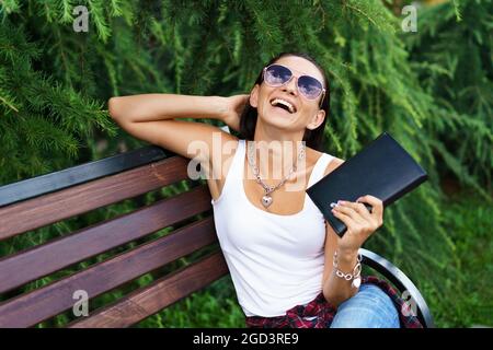 Allegra donna bruna in abiti casual e occhiali da sole si siede su una panchina di legno nel parco contro uno sfondo di cespugli verdi tiene un notebook nero in mano e sorride Foto Stock