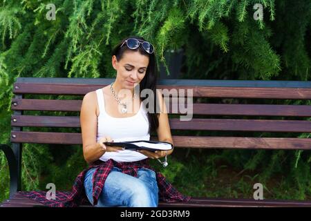 Allegra donna bruna in abiti casual e occhiali da sole si siede su una panchina di legno nel parco contro uno sfondo di cespugli verdi tiene un notebook nero in mano e sorride Foto Stock