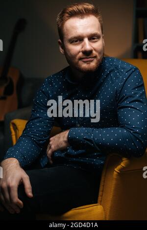 Ritratto verticale di un uomo bearded che indossa una camicia elegante seduto comodamente in una lussuosa poltrona gialla Foto Stock